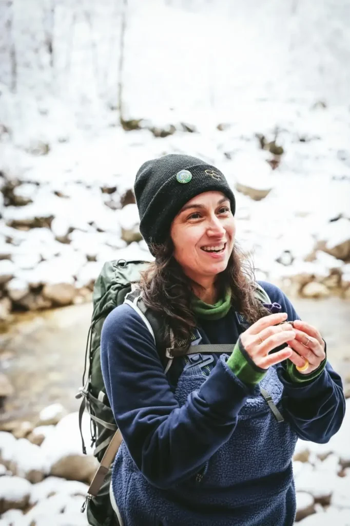 A woman with long hair, dressed warmly, standing in a snowy environment, smiling as she engages with nature.